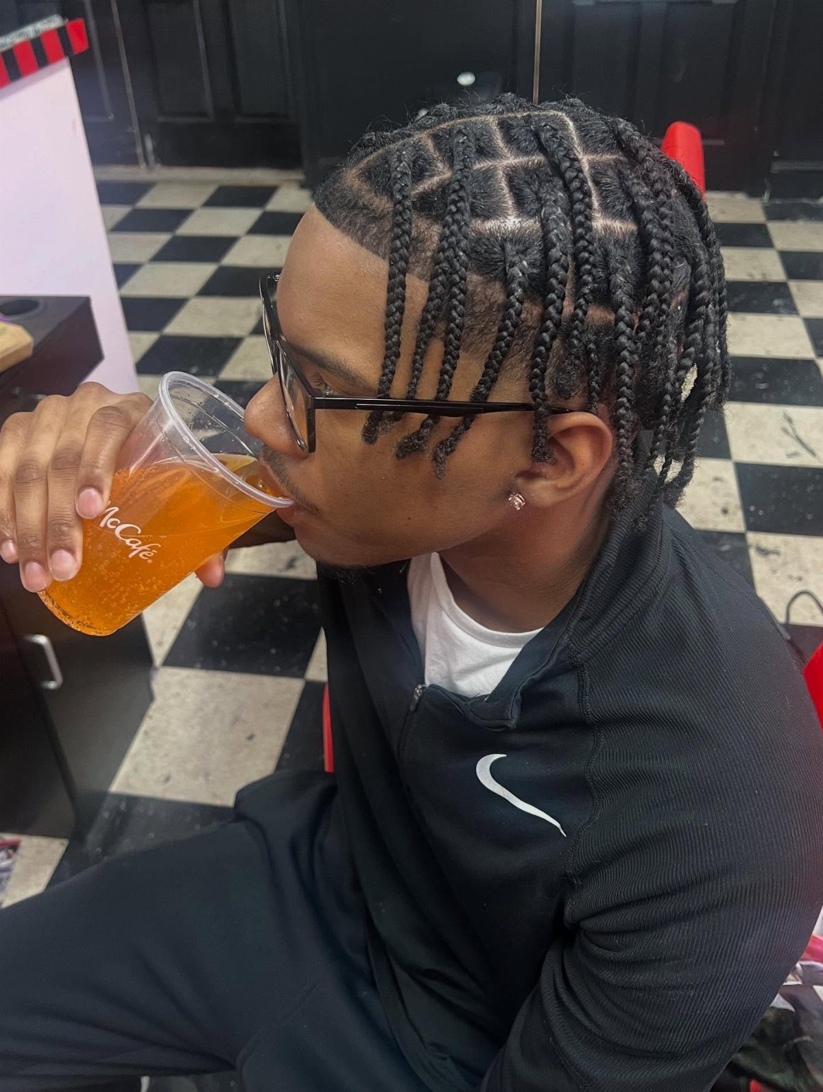 Man with square-sectioned box braids drinking from a plastic cup in a barbershop.