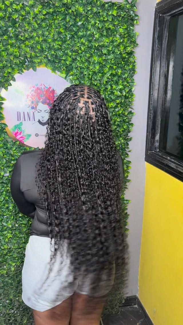 Woman with long dark boho braids featuring loose curly ends against a greenery wall