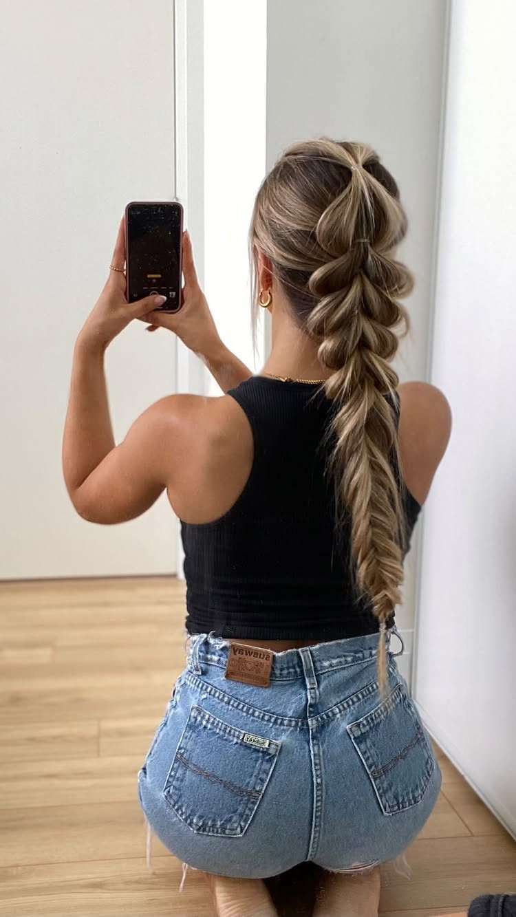 Blonde woman with a thick fishtail braid wearing a black top and denim shorts.