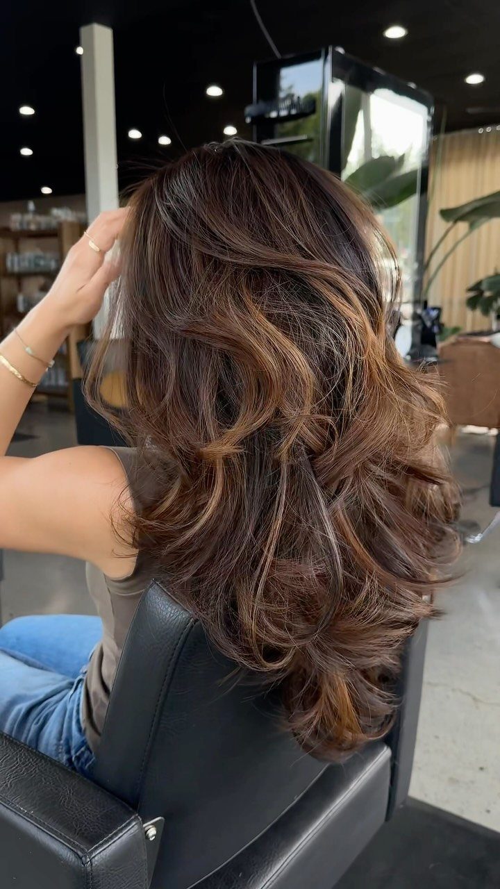 Long wavy dark brown hair with warm caramel balayage highlights in a salon.