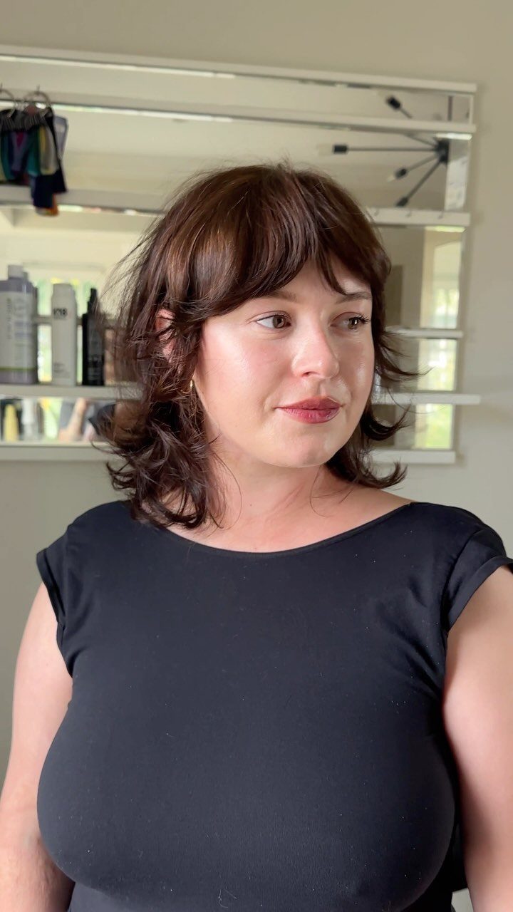 Woman with a medium length brown shag haircut and bangs wearing black top.