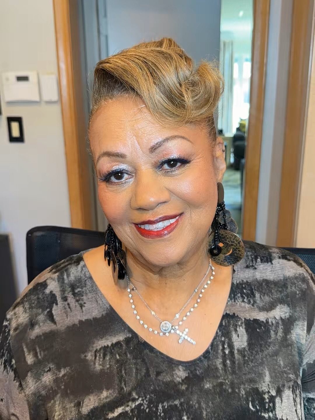 Mature woman with a blonde pompadour updo, wearing grey patterned clothing and jewelry.